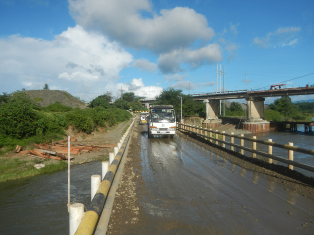 Ein Lastwagen fährt auf einer überfluteten Straße neben einer Brücke mit Geländern, Bäumen, Pfählen und einem bewölkten Himmel.