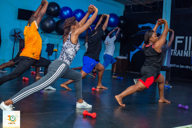 Gruppe von Menschen, die Yoga in einem Fitnessstudio praktizieren, mit Hanteln auf dem Boden, einem Banner mit der Aufschrift "Fitness-Training in Ghana" im Hintergrund und diversen Gegenständen wie Ballons, einem Ventilator und einem Stuhl.