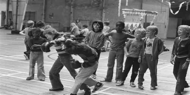 Eine Gruppe von Kindern spielt auf einem Basketballfeld in Schwarz-Weiß, mit zwei kämpfenden Personen in der Mitte und anderen auf Bänken im Hintergrund.