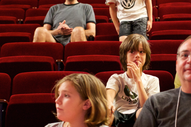 Eine Gruppe von Menschen in einem Kino mit roten Stühlen, mit einem Jungen in einem weißen T-Shirt und schwarzen Hosen in der Mitte.