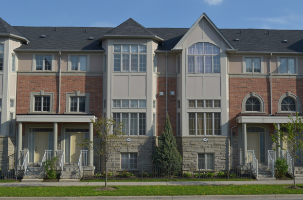 Eine Reihe von Stadthäusern in einer Wohngegend mit sichtbaren Fenstern, Türen, Säulen, Geländern, Stufen, Pflanzen, Bäumen, Gras, einer Straße im Vordergrund und dem Himmel im Hintergrund.