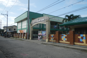 Eine Schule mit einem grünen Dach an einer Straßenecke, umgeben von Fahrzeugen, Werbetafeln, Strommasten, Kabeln, Bäumen und einem bewölkten Himmel.