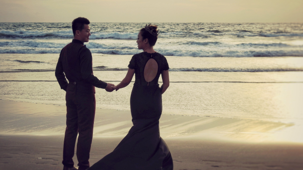 Ein Brautpaar hält sich an einem Strand bei Sonnenuntergang an den Händen, vor einem Himmel aus Orangen, Pinks und Purpurs mit tiefblauem Wasser.