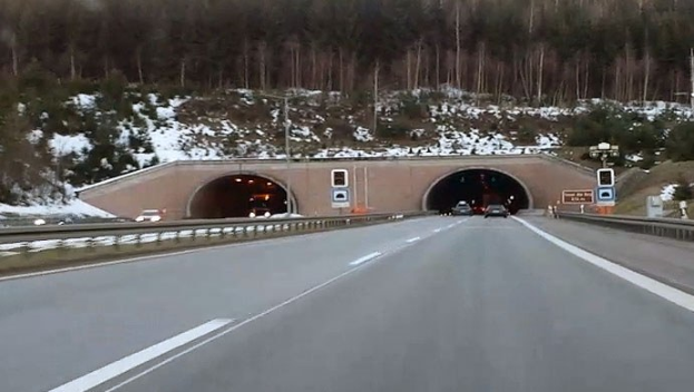 Autobahn mit Autos, die durch einen von Geländern und Schildern gesäumten Tunnel fahren, Schnee am Boden, Bäume und Masten im Hintergrund und sichtbarer Himmel darüber.