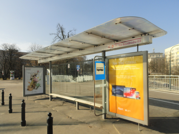 Eine Bushaltestelle mit einem Unterstand an der Straße, umgeben von Pollern, einem Zaun, Bäumen, Gebäuden und einem klaren blauen Himmel.