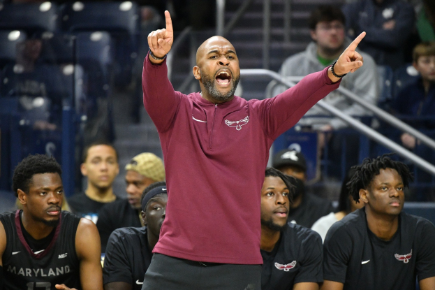 Cheftrainer des Maryland-Männer-Basketballteams in einem roten Hemd und schwarzen Shorts, der energisch gestikulierend auf der Seitenlinie eines Basketballfelds steht, umgeben von sitzenden und stehenden Personen.