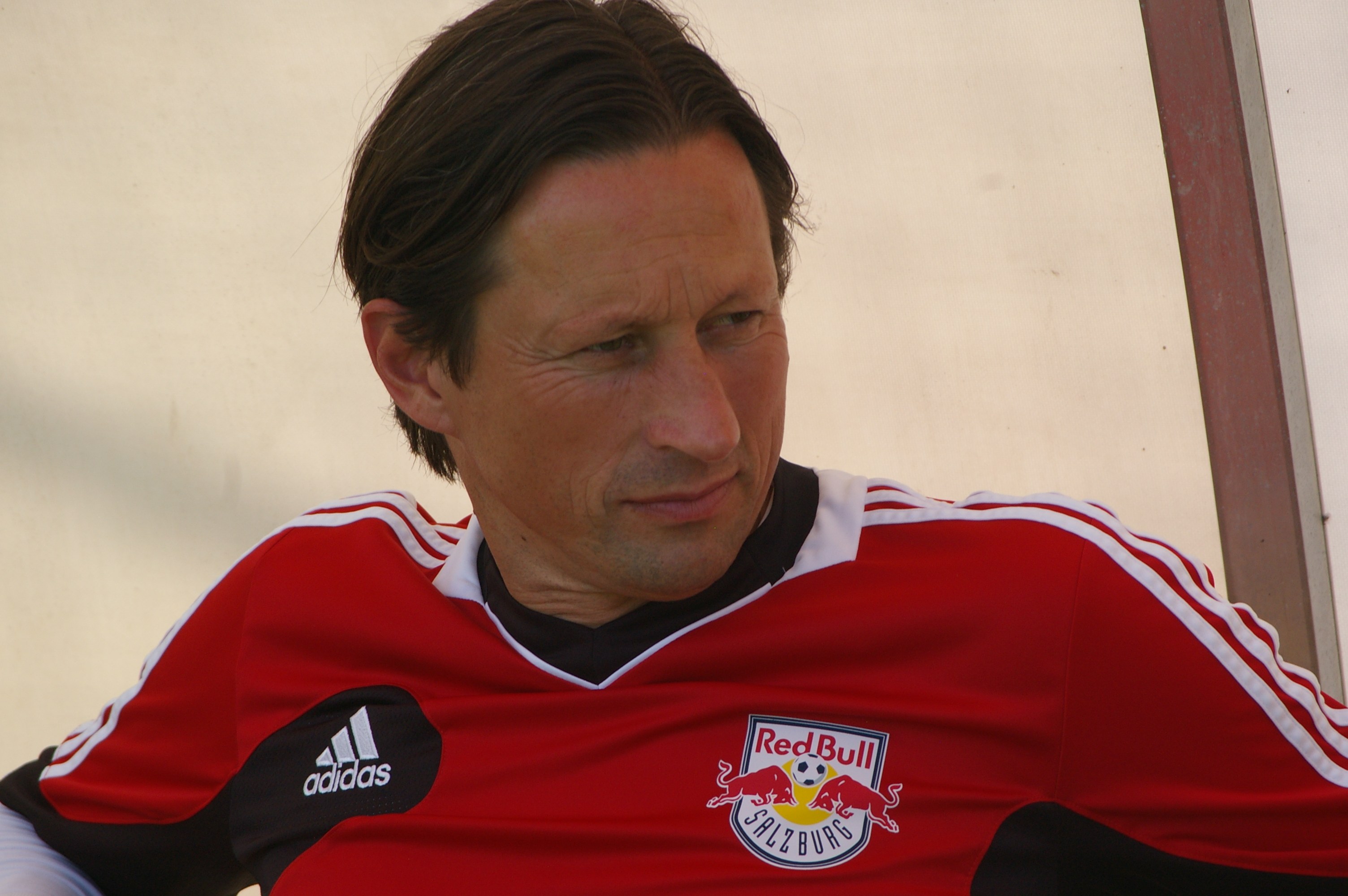 Ein professioneller Fußballspieler in einem roten und schwarzen T-Shirt sitzt auf einer Bank mit entschlossener Miene.