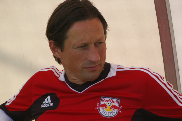 Ein professioneller Fußballspieler in einem roten und schwarzen T-Shirt sitzt auf einer Bank mit entschlossener Miene.