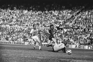 Schwarzes und weißes Foto von Männern, die auf einem Feld Fußball spielen, mit Zuschauern im Hintergrund und einem Banner 'Bundesliga 1978-79 Ajax 1-1 Hannover 96'.