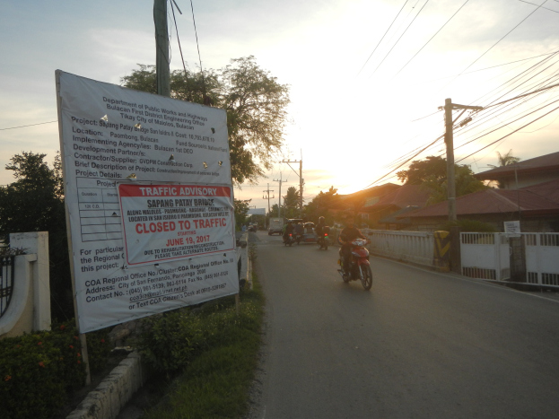 Eine Gruppe von Motorradfahrern auf einer Straße mit einem "Verkehrshinweis Geschlossen für den Verkehr"-Schild, umgeben von einer blühenden Wand, Bäumen, Strommasten, Häusern und einem klaren blauen Himmel.