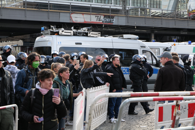 Eine Gruppe von Menschen steht in der Nähe von Polizeifahrzeugen mit Barrieren im Vordergrund, einige tragen Helme und halten Telefone, mit einer Brücke und Gebäuden im Hintergrund während einer Demonstration in Berlin.