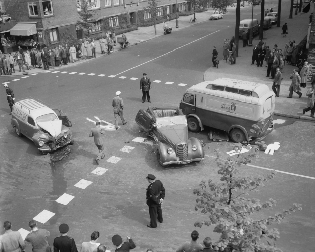 Schwarz-weißes Bild einer Autounfall-Szene an einer Straßenkreuzung mit einem Van, anderen Fahrzeugen und einer Gruppe von Menschen in der Nähe, umgeben von Gebäuden, Bäumen, Pfählen und Texttafeln.