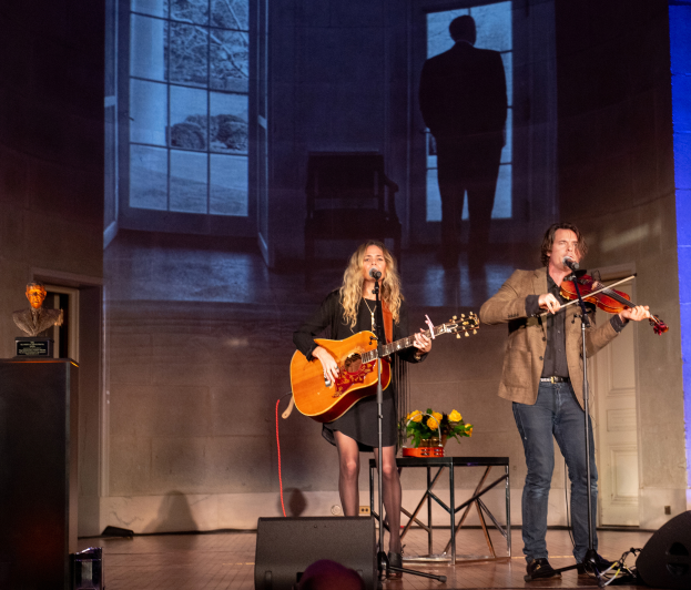 Ein Mann in einer braunen Jacke und blauen Hosen sowie eine Frau mit einer Gitarre und einem Mikrofon singend auf der Bühne vor einem Publikum, mit einer Vase auf dem Tisch zwischen ihnen und Lautsprechern am unteren Ende der Bühne und einer Wand mit einem Bildschirm im Hintergrund.