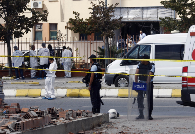 Eine Gruppe von Polizeibeamten steht vor einem Fenster mit Fenstern, umgeben von Fahrzeugen, einem Zaun, Bäumen und Trömmern, darunter Ziegelsteine und ein Helm auf dem Boden, wahrscheinlich nach einer Bombenexplosion in Mexiko-Stadt.