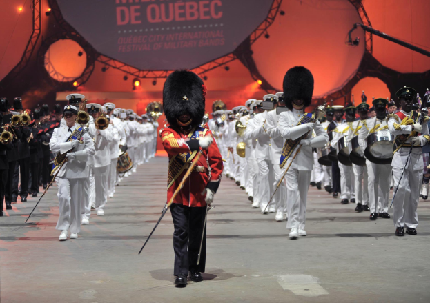 Gruppe uniformierter Musiker marschiert in Formation, einige spielen Instrumente, mit einer beleuchteten Tafel mit Text im Hintergrund während eines Ereignisses.