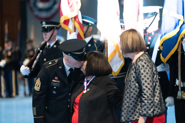 Ein Mann und eine Frau küssen sich, während sie sich in Gegenwart einer Gruppe uniformierter Personen mit Fahnen umarmen.