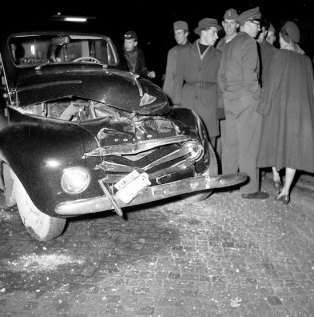 Ein Schwarz-Weiß-Bild eines verunglückten Autos am Straßenrand, dessen Motorhaube eingedrückt und Fenster zersplittert sind, umgeben von einer Gruppe besorgter Menschen.