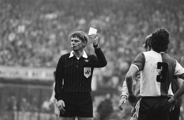 Ein Schiedsrichter in Schwarz und Weiß hält eine rote Karte vor einer Menge in Fußballtrikots, mit einem Zaun im Hintergrund.