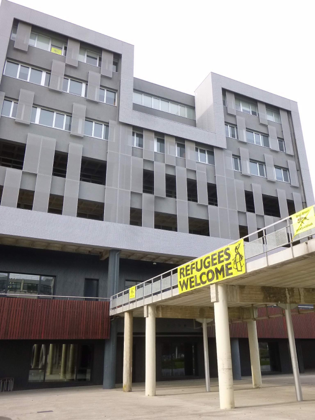 Großes Gebäude mit einer "Flüchtlinge Willkommen"-Schrifttafel, Säulen, Geländer und Himmel im Hintergrund.