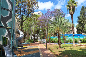 Spielplatz mit einer Rutsche, Palmen, Gebäuden, Straßenlaternen, Grills, Pflanzen, Bäumen, Gras, Felsen und einem bewölkten Himmel im Hintergrund.