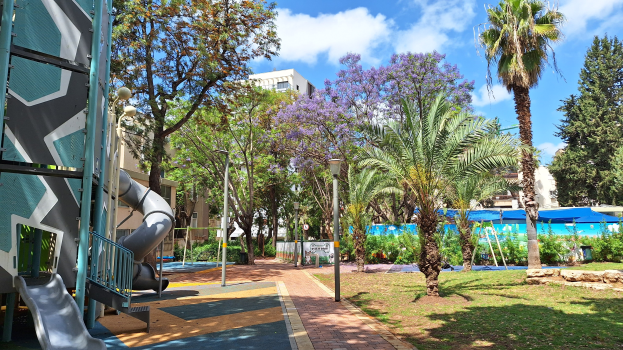 Spielplatz mit einer Rutsche, Palmen, Gebäuden, Straßenlaternen, Grills, Pflanzen, Bäumen, Gras, Felsen und einem bewölkten Himmel im Hintergrund.