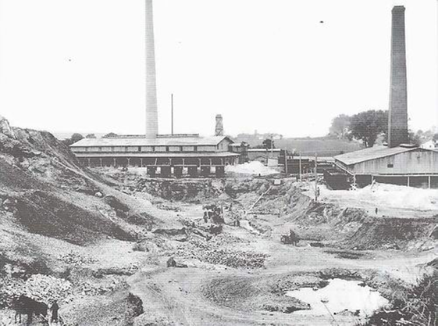 Ein Schwarz-Weiß-Foto einer Baustelle mit zwei Schornsteinen im Hintergrund, umgeben von Gebäuden, Masten, Bäumen und einem klaren Himmel, mit Text unten, der darauf hinweist, dass es ein Steinkohlekraftwerk ist.