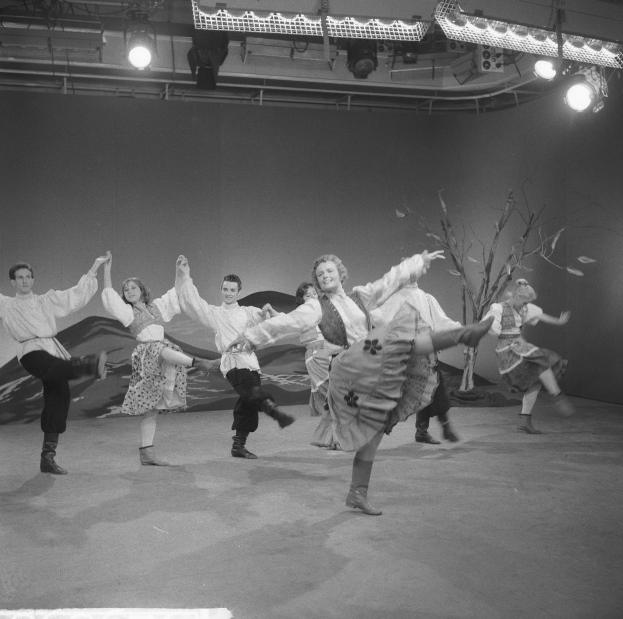 Schwarzes und weißes Bild einer Gruppe von Menschen, die auf einer Bühne tanzen, mit Deckenleuchten und einer Wand im Hintergrund.