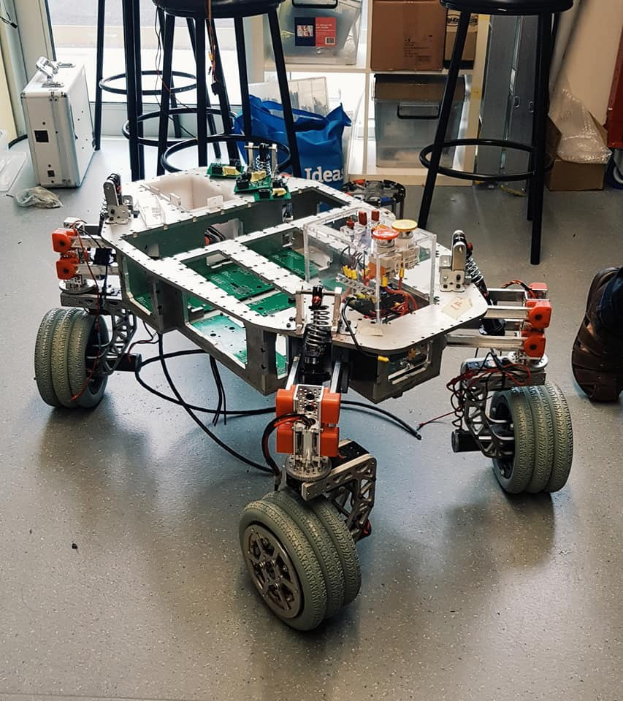 Ein Roboter sitzt auf einem Tisch in einem Raum mit Hockern, Kartons und einem sichtbaren Bein einer Person auf der rechten Seite.