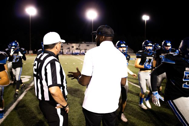 Ein Fußballtrainer in einer Mütze spricht mit seinem Team auf dem Feld bei Nacht, mit Spielern in Helmen, Laternenmasten und einem dunklen Himmel im Hintergrund.