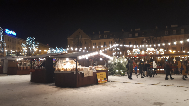 Ein nächtlicher Weihnachtsmarkt auf einer verschneiten Straße mit Menschen, Ständen, Pflanzen, Gebäuden und Schildern unter einem bewölkten Himmel.