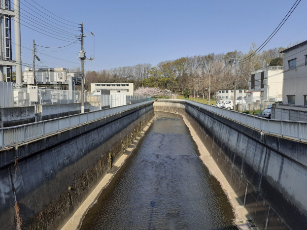 Ein kleiner Kanal verläuft durch eine Stadt mit Gebäuden auf beiden Seiten, Strommasten mit Kabeln und Fahrzeugen auf der Straße, mit Bäumen und einem klaren blauen Himmel im Hintergrund.