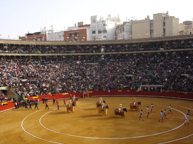 Menschen gehen auf dem Boden, einige reiten Pferde, andere sitzen im Stadion und schauen zu, umgeben von Fahnen, Gebäuden und dem Himmel.