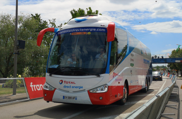 Ein roter und weißer Bus fährt auf einer Straße neben einem Zaun, mit Passagieren im Inneren; auf der rechten Seite befindet sich ein Geländer, und im Hintergrund sind Bäume, Pfosten, Verkehrszeichen, eine Brücke und ein bewölkter Himmel zu sehen.
