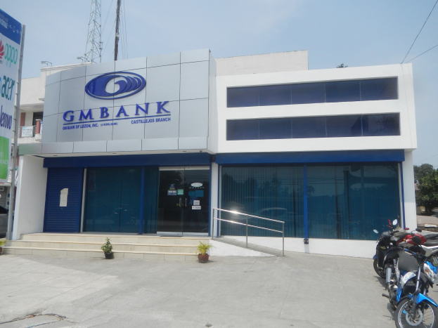Außenansicht der Gmbank Bank in Hyderabad mit Glaswänden, Treppenstufen mit Geländern, Topfpflanzen, einem Banner, nahestehenden Fahrzeugen, einem Versorgungsturm mit Drähten, Bäumen und einem bewölkten Himmel.