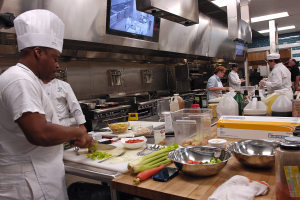 Eine Gruppe von Köchen in einer Küche beim Zubereiten von Speisen, mit einem in weißer Uniform und Mütze mit einem Messer, umgeben von Utensilien, Gemüse und anderen Gegenständen auf einem Schneidbrett.