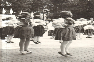 Schwarzes und weißes Foto von Frauen in traditioneller bayrischer Tracht, die auf einem Holzfußboden tanzen, umgeben von Bäumen und Fahnen.