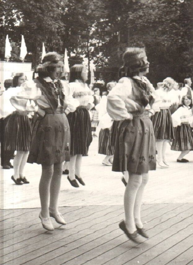 Schwarzes und weißes Foto von Frauen in traditioneller bayrischer Tracht, die auf einem Holzfußboden tanzen, umgeben von Bäumen und Fahnen.