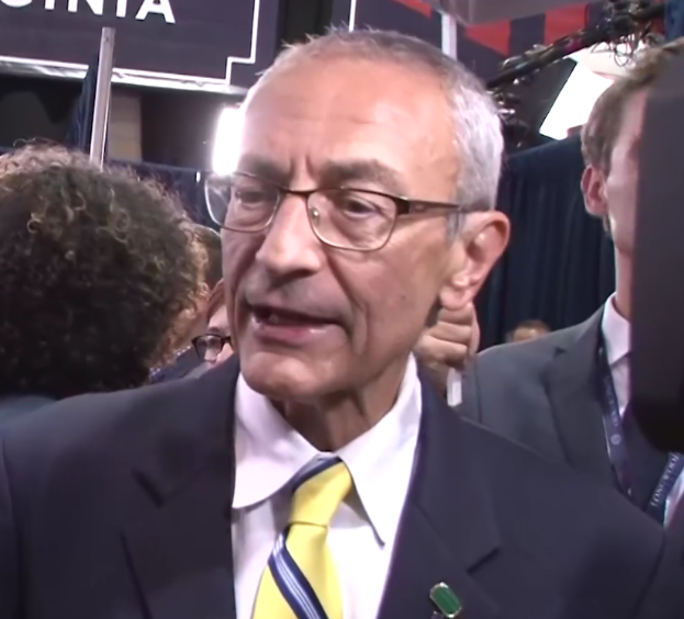 Ein Mann im Anzug und mit Krawatte, der eine Brille trägt, spricht zu Reportern mit einem Mikrofonständer vor sich, umgeben von einer Gruppe von Menschen, mit Scheinwerfern und einer Tafel im Hintergrund.