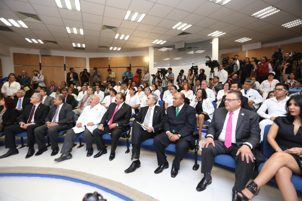 Eine große Gruppe von Menschen, die auf Stühlen in einem Raum sitzen, einige halten Kameras, mit einer Wand, einer Decke und einem Boden im Hintergrund.