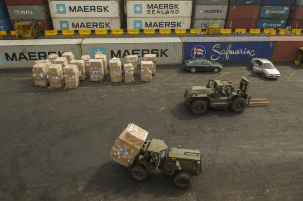 Militärfahrzeug vor einem großen Container Schiff mit mehreren Fahrzeugen und verstreuten Kartonagen auf der Straße und textbehafteten Containern im Hintergrund.
