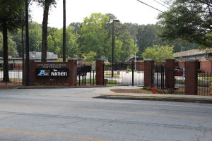 Einfahrt zu einer Schule mit einem Schild, umgeben von B├Ąumen, Stra├čenm├Âbeln, Fahrzeugen und Geb├Ąuden unter einem klaren blauen Himmel.