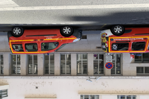 Zwei Krankenwagen nebeneinander in einer Garage geparkt mit einem Gebäude mit Glasfenstern und Schildern im Hintergrund und einer Überwachungskamera an der Wand unten im Bild.