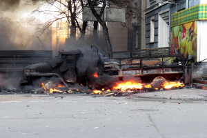 Ein von Flammen umgebener Lkw am Rande einer Straße, mit Rauch, der aus ihm aufsteigt, vor dem Hintergrund von Bäumen, Gebäuden und einem klaren Himmel.