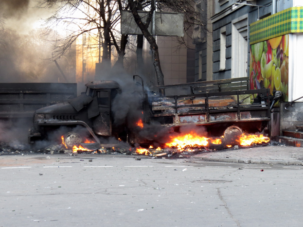Ein von Flammen umgebener Lkw am Rande einer Straße, mit Rauch, der aus ihm aufsteigt, vor dem Hintergrund von Bäumen, Gebäuden und einem klaren Himmel.
