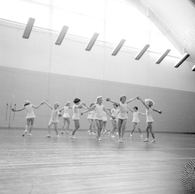 Schwarzes und weißes Bild von jungen Mädchen, die in einem Studio tanzen und sich an den Händen halten, mit einem Ball auf der rechten Seite und Lampen an der Decke.