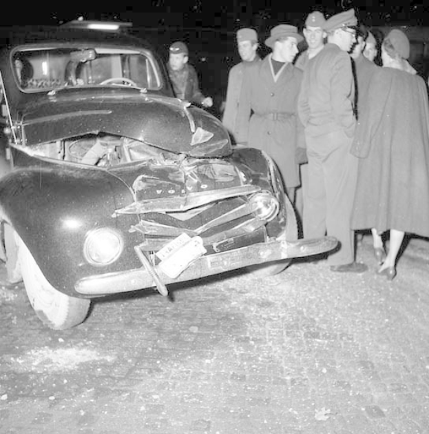 Schwarz-weißes Bild eines schwer beschädigten Autos am Straßenrand mit einer Gruppe von Menschen, die es begutachten.