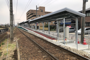 Ein Bahnhof mit einem Zug auf den Gleisen, umgeben von Gebäuden, Strommasten, Stromkabeln, Kraftfahrzeugen, Verkehrskegeln, Informationsanzeigen, Zäunen, Bäumen und einem klaren blauen Himmel.