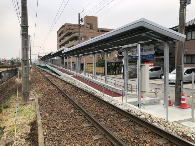 Ein Bahnhof mit einem Zug auf den Gleisen, umgeben von Gebäuden, Strommasten, Stromkabeln, Kraftfahrzeugen, Verkehrskegeln, Informationsanzeigen, Zäunen, Bäumen und einem klaren blauen Himmel.