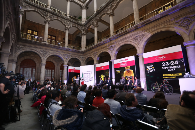Eine Gruppe von Menschen, die an einem Tisch mit einem Podium und einem Mikrofon vor einer Menge, Kameras und Bannern sitzen, was eine Pressekonferenz für das CDMX 2017-Event vermuten lässt.