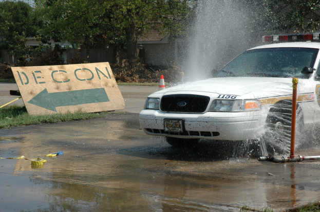 Polizeiauto fährt durch eine Pfütze mit verstreuten Gegenständen, darunter eine Platte, ein Verkehrskegel und andere Objekte; Bäume, Gebäude und ein klarer blauer Himmel im Hintergrund.